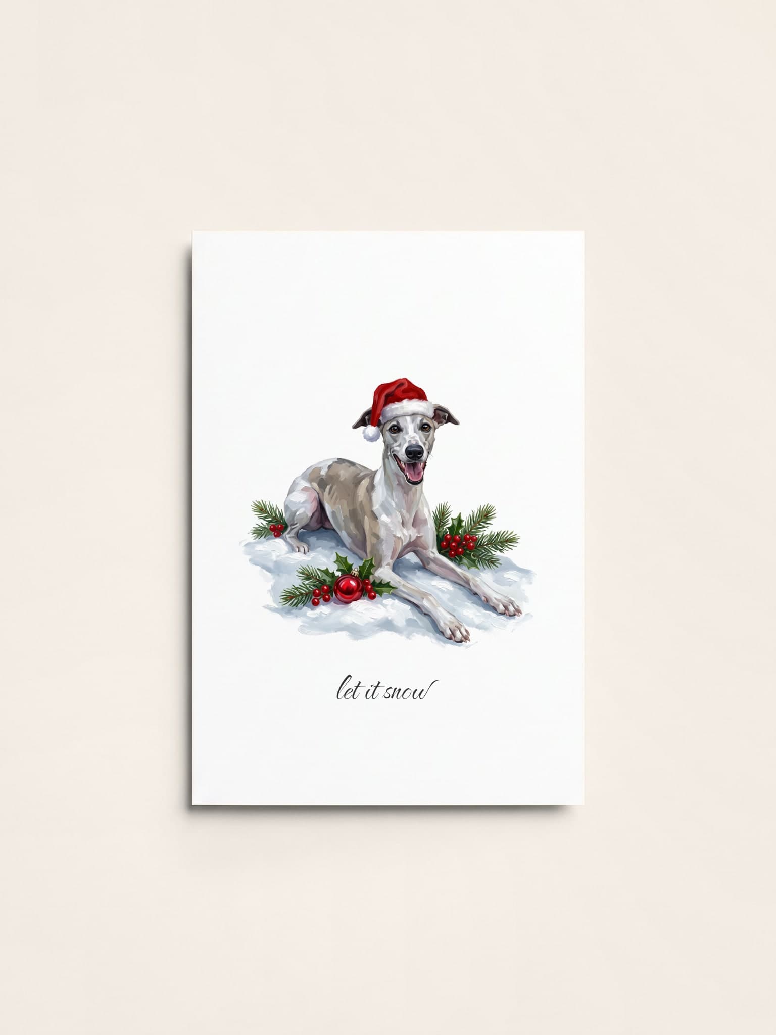 A Christmas card of a cream whippet in a Santa hat lying in the snow with holly and a red bauble